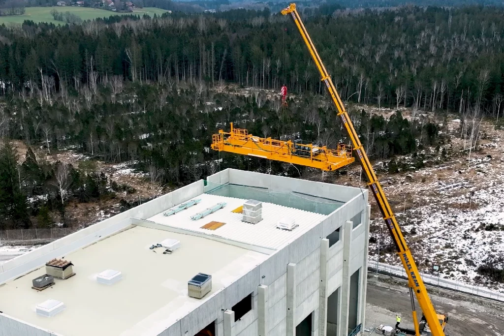 Ein orangener Kran hebt den automatisierten Beschickungskran durch das Dach einer Biomasseanlage in Bayern. Im Hintergrund der Biomasseanlage ist ein Wald zu erkennen.
