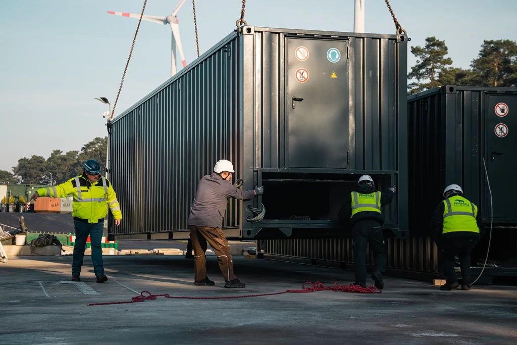 Bauarbeiter schieben an Kran und Ketten befestigten schwarzen Container in Steinfurt. Drei von den vier Männer tragen gelbe Schutzwesten. Im Hintergrund kann man ein Windrad erkennnen.