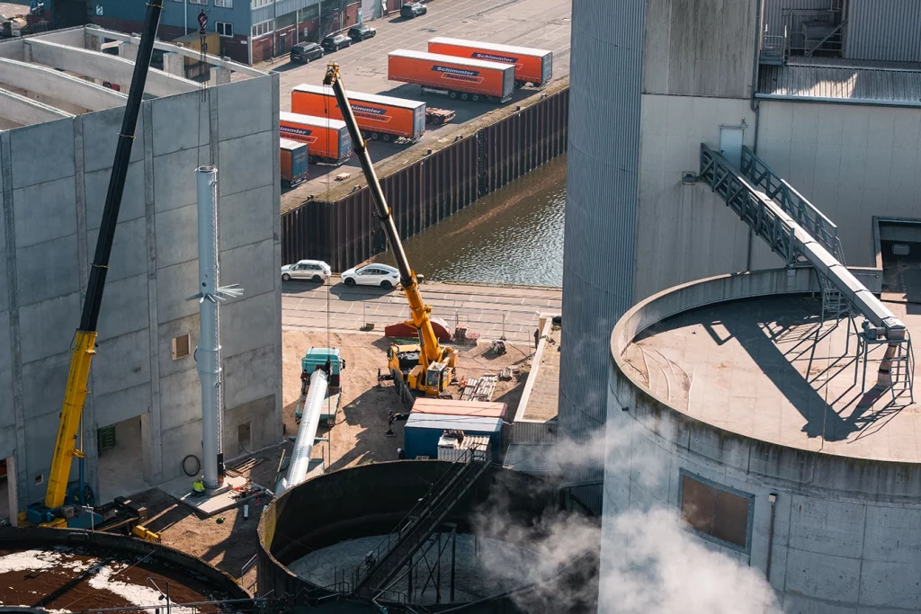 Drohnenaufnahme von der Biomasseanlage bei Avangard in Bremen. Ein Kran setz einen Schornstein in den Boden. Ein anderer, noch liegender Schornstein ist befestigt an einem zweiten Kran.