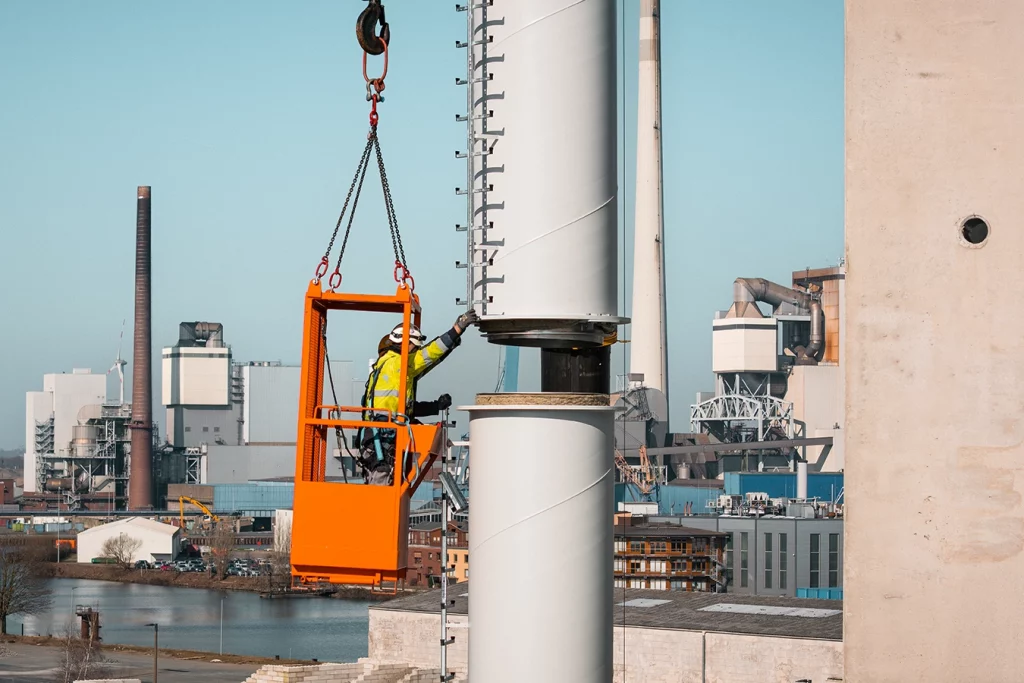 Ein Teil eines Schornsteins wird auf einen anderen Teil gesetzt. Zwei Mitarbeiter in einem Art kleinen orangefarbigen Aufzug, welcher von einem Kran gehalten wird, kontrollieren das Einsetzen.