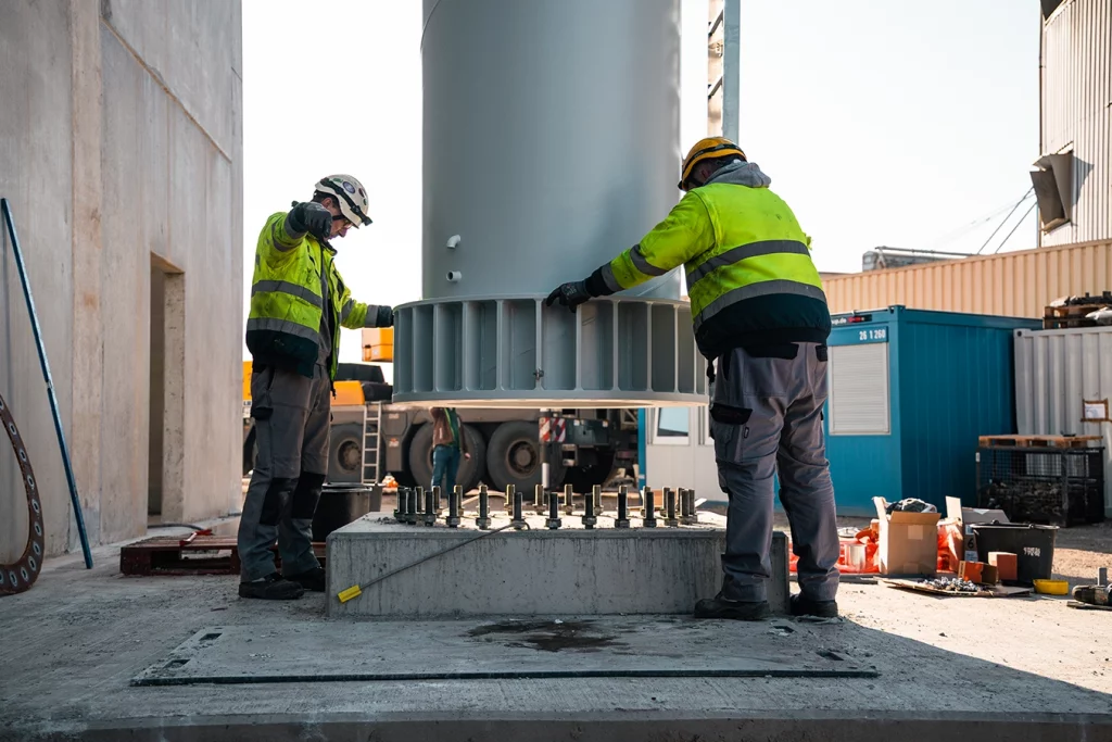 Zwei Bauarbeiter mit gelber Sicherheitsweste kontrollieren das Einsetzen eines Schornsteins in den Boden.