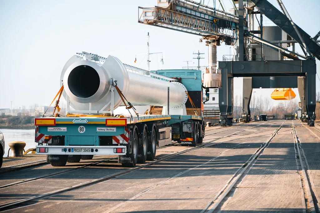 Ein türkisfarbener LKW transportiert ein Teil eines weißen Schornsteins. Die Szene spielt sich am Hafen in Bremen ab. Die Weser liegt linksseits vom LKW.