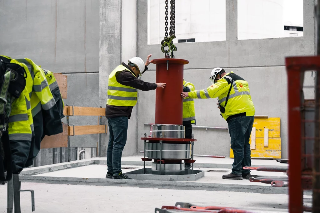 Ein Kran lässt die Voreinrichtung einer Turbine hinunter und setzt sie in den Boden einer Biomasseanlage ein. Drei Mitarbeiter mit gelber Sicherheitsweste kontrollieren dabei den Vorgang. Sie stehen an der Voreinrichtung und berühren diese.