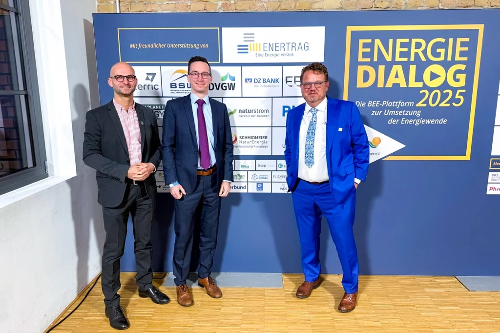 Thomas Schmidmeier, Gerrit Kettel und eine weitere Person stehen vor einer Sponsorentafel des Energiedialogs 2025