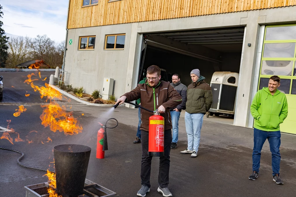 Ein Mitarbeiter von Schmidmeier NaturEnergie GmbH löscht mit einem Feuerlöscher ein kleines Feuer. Um ihn herum stehen vier weitere Mitarbeiter und betrachten die Situation. Die Personen befinden sich draußen auf dem Firmengelände von Schmidmeier NaturEnergie in Ödenthal.