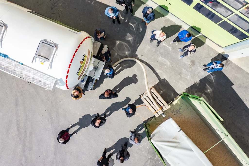 Eine Gruppe von Teilnehmenden der DEPI Fahrerschulung stehen um einen LKW und Anhänger herum. Sie schauen zu wie Hackschnitzel durch einen Schlauch von dem LKW in den Anhänger gelangen. Die Szene sieht man aus der Vogelperspektive.