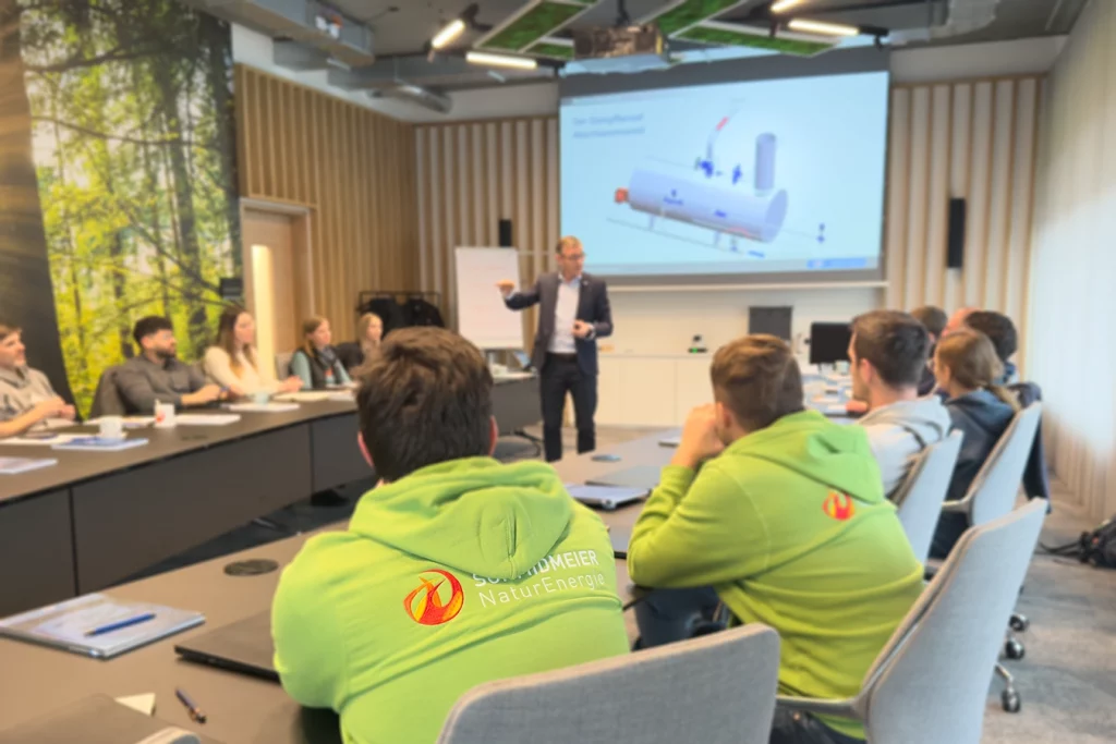 Ein Mann referiert vor einer Leinwand, um ihn herum sitzen an zwei länglichen und parallel verlaufenden Tischen die Teilnehmer der Weiterbildung.