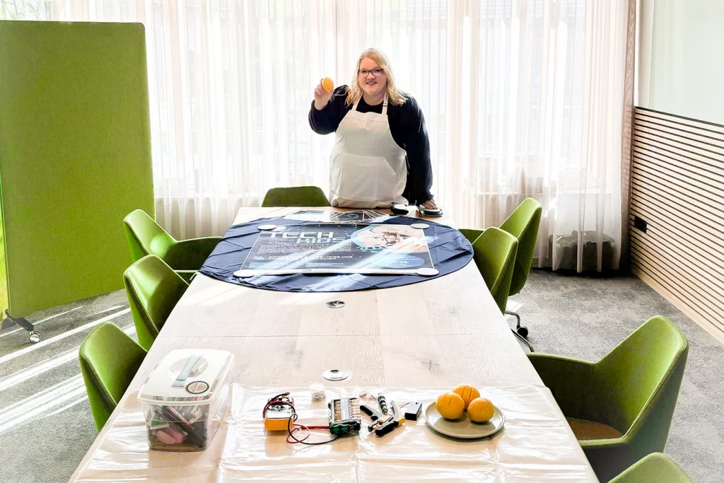 Frau steht hinter einem länglichen Tisch in einem sonnen-beleuchteten Raum mit grünen Stühlen. Die Frau hält eine Zitrone in der Hand. Auf dem Tisch befinden sich weitere Zitronen und technische Sachen sowie Stifte, die für die Kinder zum Basteln bereitstehen.