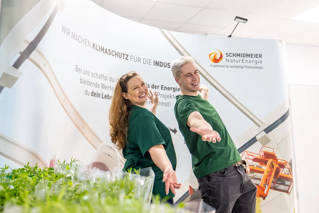 Eine Mitarbeiterin und ein Mitarbeiter von Schmidmeier NaturEnergie stehen am Messestand und breite ihre Arme aus. Hinter ihnen befindet sich der Messebanner mit Schmidmeier Logo.