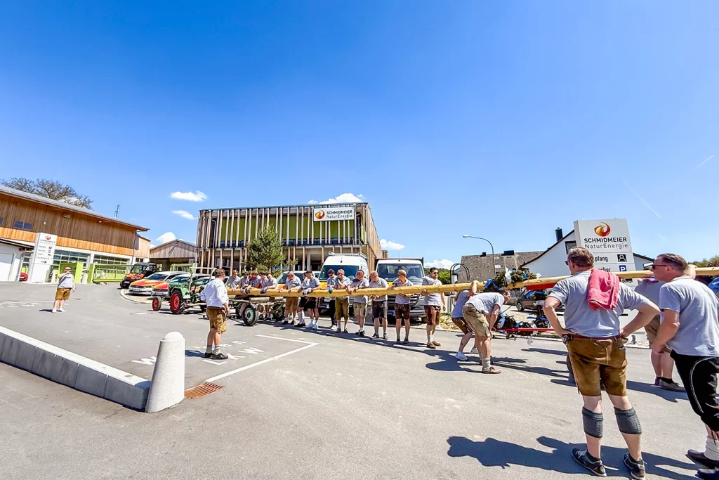 Eine Gruppe von Männern in Tracht transportiert bei schönem und klarem Wetter einen Maibaum. Im Hintergrund erkennt man das Firmengebäude der Schmidmeier NaturEnergie.