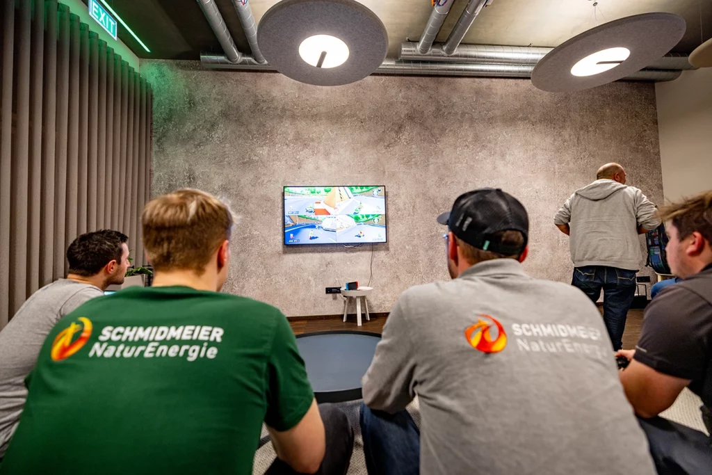Vier Schmidmeier NaturEnergie Mitarbeiter sitzen auf einer Couch und spielen auf dem an der Wand hängenden Fernseher das Videospiel Mario Kart. Vor ihnen steht ein schwarzer runder Tisch. Die Wand ist grau.