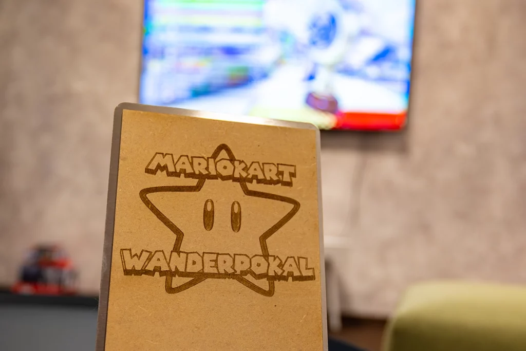 Ein Pokal ist im Vordergrund vor einem laufende Fernseher zu sehen. Auf dem Pokal ist der Stern, der im Videospiel Mario Kart eine wichtige Rolle spielt, abgebildet. Über dem Stern steht "Mario Kart", darunter "Wanderpokal".