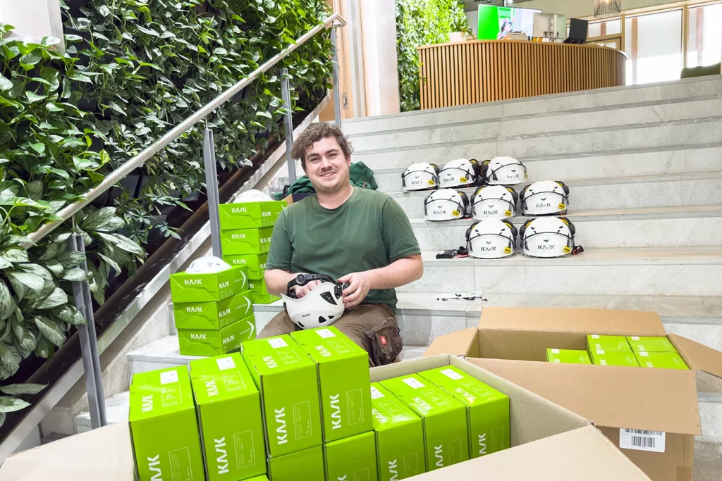 Mitarbeiter Philipp Hierl von der Schmidmeier NaturEnergie GmbH sitzt auf der Treppe des Eingangsbereichs. Auf seinen Oberschenkeln liegt ein Sicherheitshelm. Um ihn herum befinden sich grüne Pakete mit Helmen sowie fertig zusammengebaute Helme.