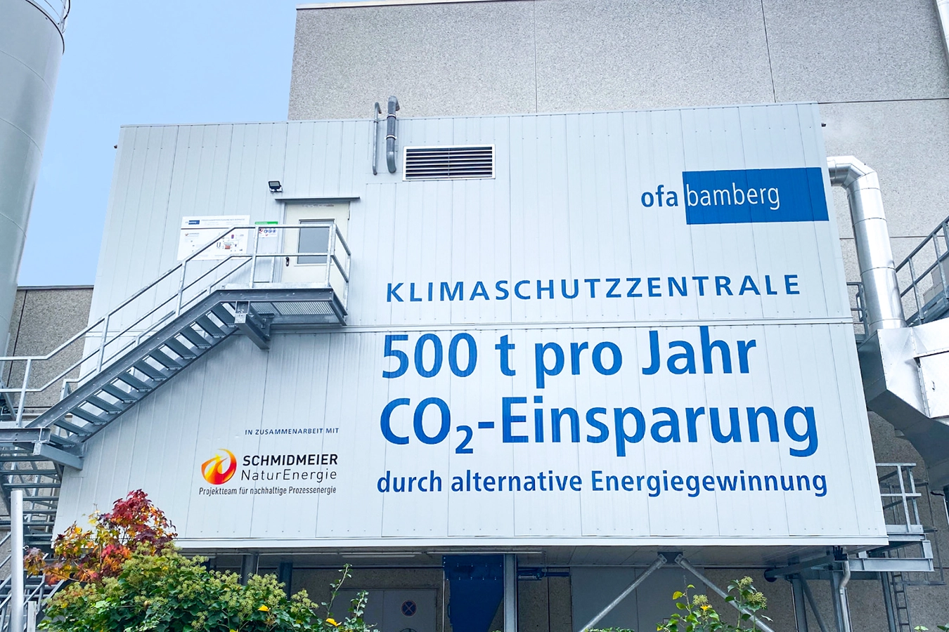 Biomassedampfanlage in Bamberg. Ein grauer rechteckiger Kasten ist rechts verbunden mit einem metallenem Rohr. Links befindet sich eine eine Treppe, die zu einer Tür führt. Die Tür befindet sich über der Hälfte des Kastens. Im Hintergrund befindet sich ein Gebäude aus Stein.