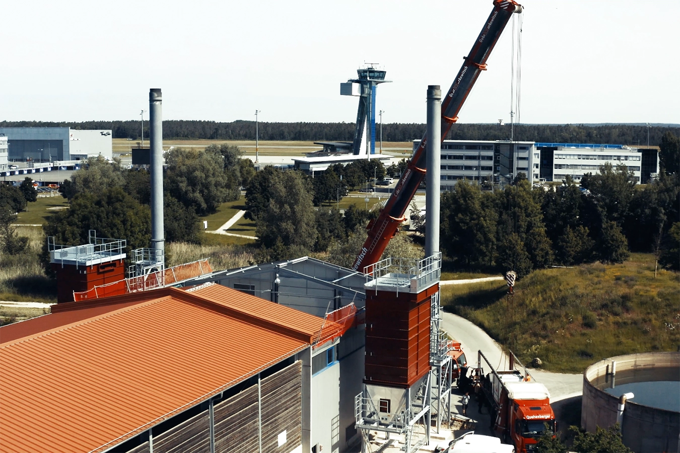 Luftaufnahme der Biomasseanlage mit doppelten Biomassekessel in Nürnberg. Das Gebäude der Anlage hat ein orangenes Dach und zwei Kamine stehen am Schluss des Gebäudes. Im Hintergrund ist ein roter Kran sowie der Nürnberger Flughafen.