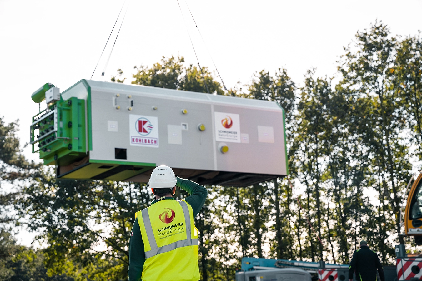 Ein Mitarbeiter der Schmidmeier NaturEnergie mit Sicherheitsweste steht vor einer Komponente, die mit einem Kran transportiert wird. Er hält sich seine Hand vor das Gesicht um sich von der Sonne zu schützen.