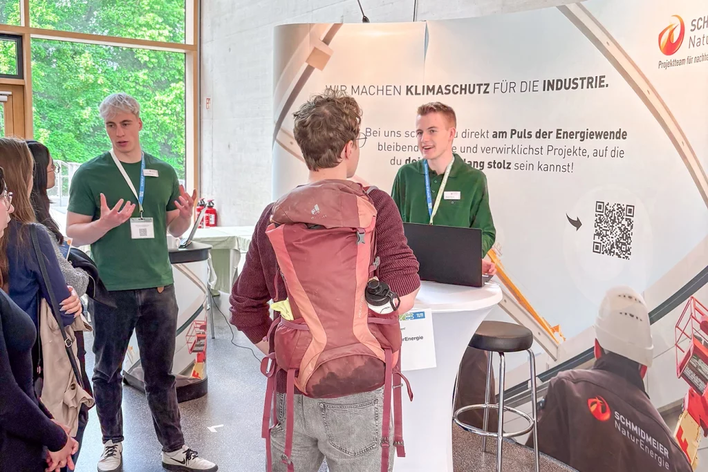 Zwei Mitarbeiter von Schmidmeier NaturEnergie stehen am Messestand und unterhalten sich mit interessierten Studenten.