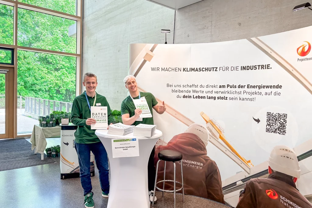 Zwei Mitarbeiter der Schmidmeier NaturEnergie stehen vor dem Messestand an der TUM in Straubing und lachen in die Kamera.