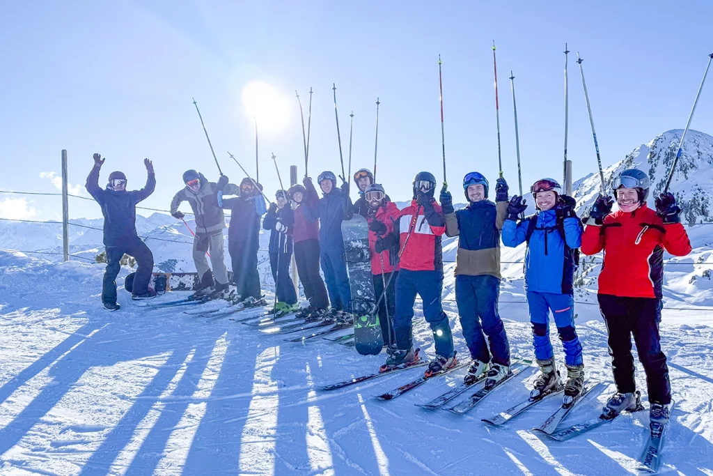 Mitarbeiter von Schmidmeier NaturEnergie GmbH stehen mit Ski aufgereiht in einer Reihe und strecken ihre Skistöcke in die Luft. Hinter ihnen befindet sich ein beschneiter Berg und die Sonne scheint.