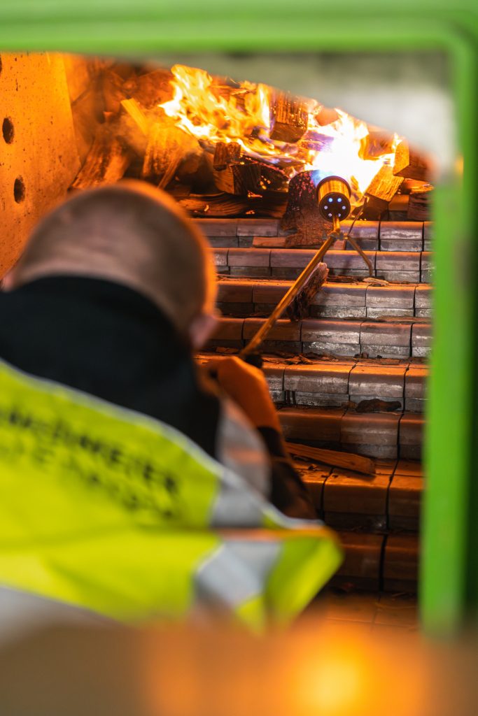 Ein Mitarbeiter verbrennt mit Hilfe eines Gasfeuerzeugs die Holzstücke im Ofen. Feuer entsteht.
