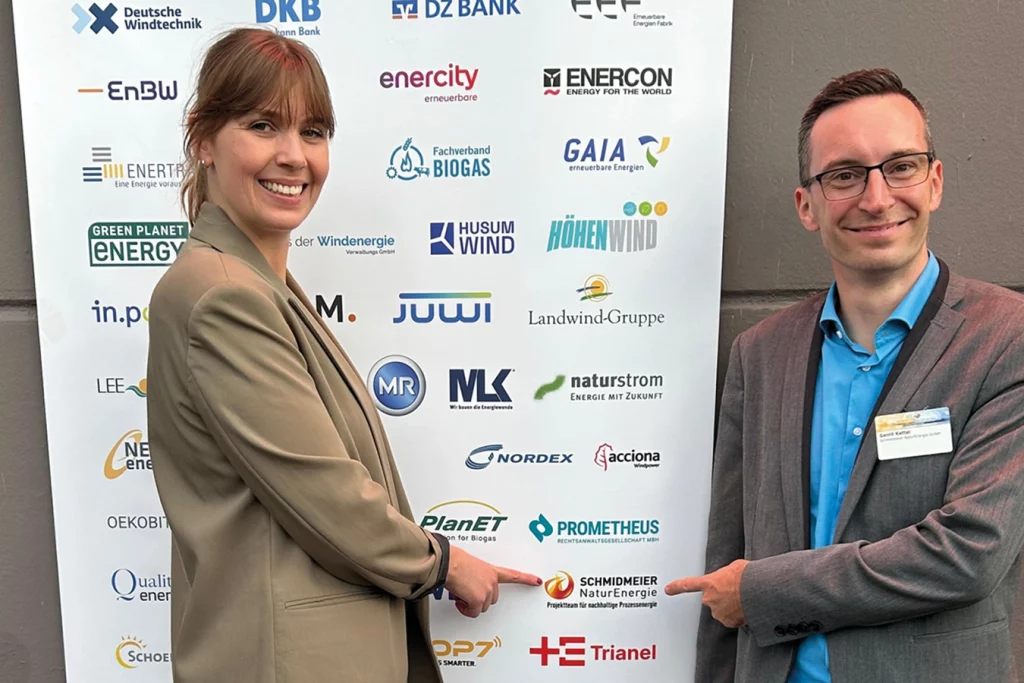 Zwei Mitarbeiter der Schmidmeier NaturEnergie stehen vor einem weißen Plakat mit Firmenlogos. Link, Anna-Marlen Vöcking und rechts, Gerrit Kettel zeigen mit ihrem Zeigefinger auf das Logo von der Schmidmeier NaturEnergie, welches sich auf dem Plakat befindet.