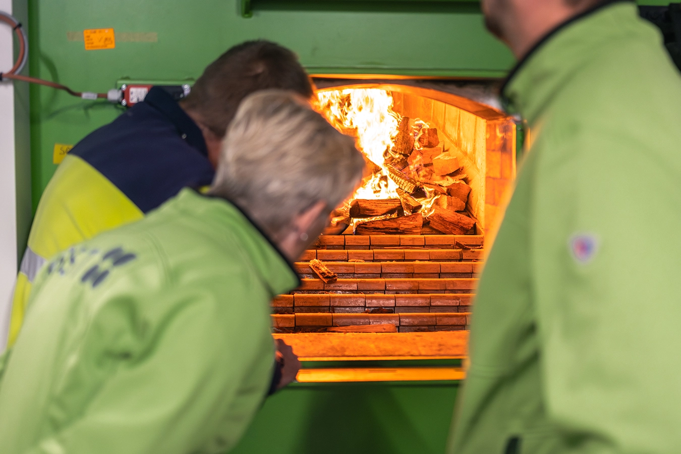 Drei Personen schauen in einen brennenden Biomassekessel. Zwei davon tragen eine grüne Jacke.