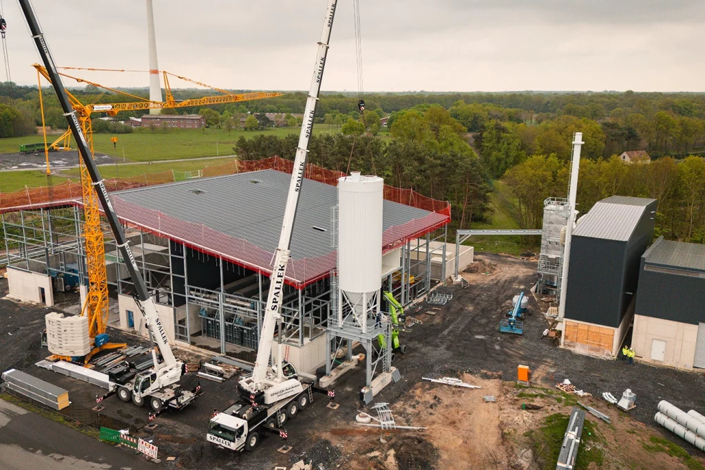 Baustelle einer Biomasse-Anlage, auf der sich Kräne, Baustellengeräte und Maschinen. Ein weißer großer Kran hebt ein Silo in seine Befestigung. Ein Silo ist ein großer Speicher für Schuttgüter, in diesem Fall der Biomasse. Im Hintergrund ist eine grüne Landschaft mit Bäumen zu sehen.