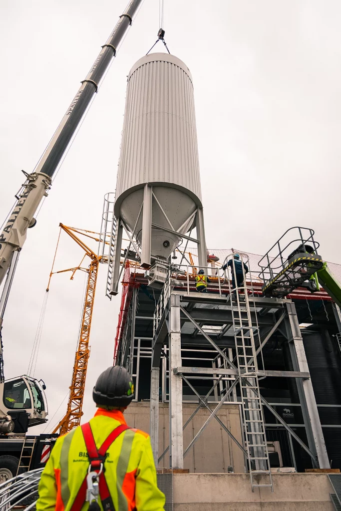 Ein weißer großer Kran hebt ein Silo in seine Befestigung. Ein Silo ist ein großer Speicher für Schuttgüter, in diesem Fall der Biomasse. Ein Bauerbeiter in neongelber Jacke beobachtet das ganze Geschehen.