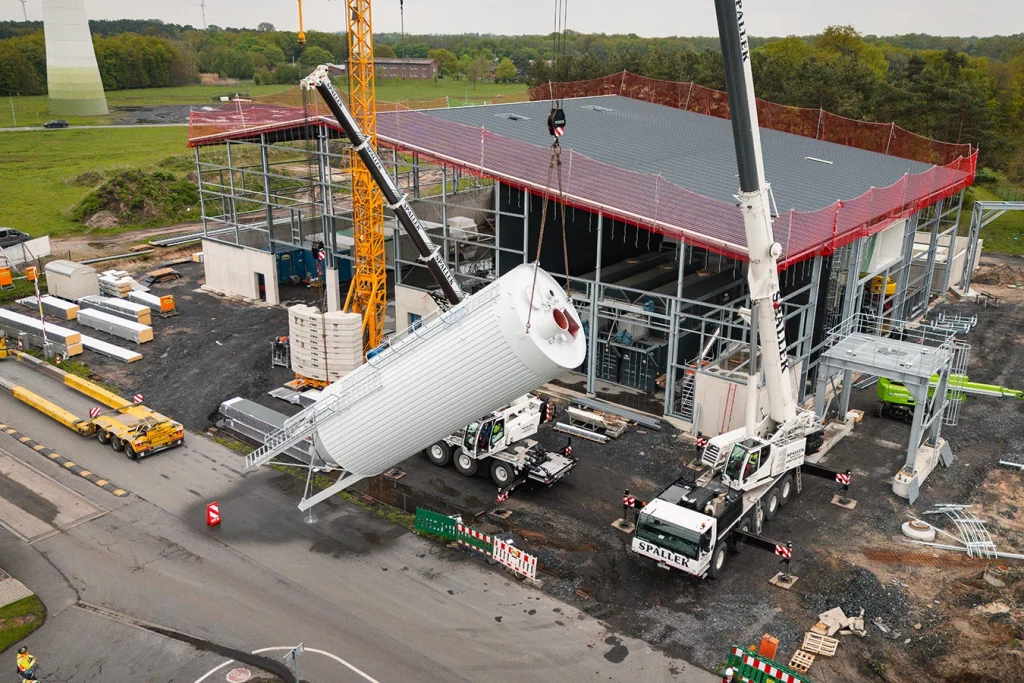 Baustelle einer Biomasse-Anlage, auf der sich Kräne, Baustellengeräte und Maschinen befinden. Ein weißer großer Kran hebt ein Silo in seine Befestigung. Ein Silo ist ein großer Speicher für Schuttgüter, in diesem Fall der Biomasse. Im Hintergrund ist eine grüne Landschaft mit Bäumen zu sehen.