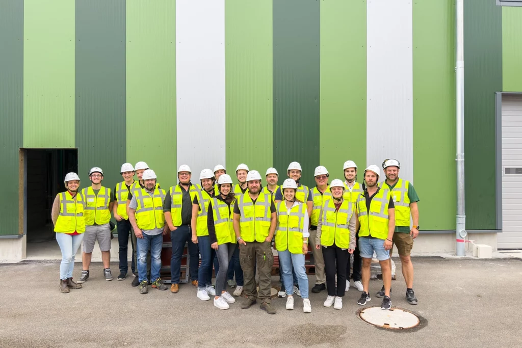 Gruppenbild (19 Personen) stehen vor einer grünen Biomasseanlage in Dingolfing. Alle tragen gelbe Warnweste und einen Sicherheitshelm.
