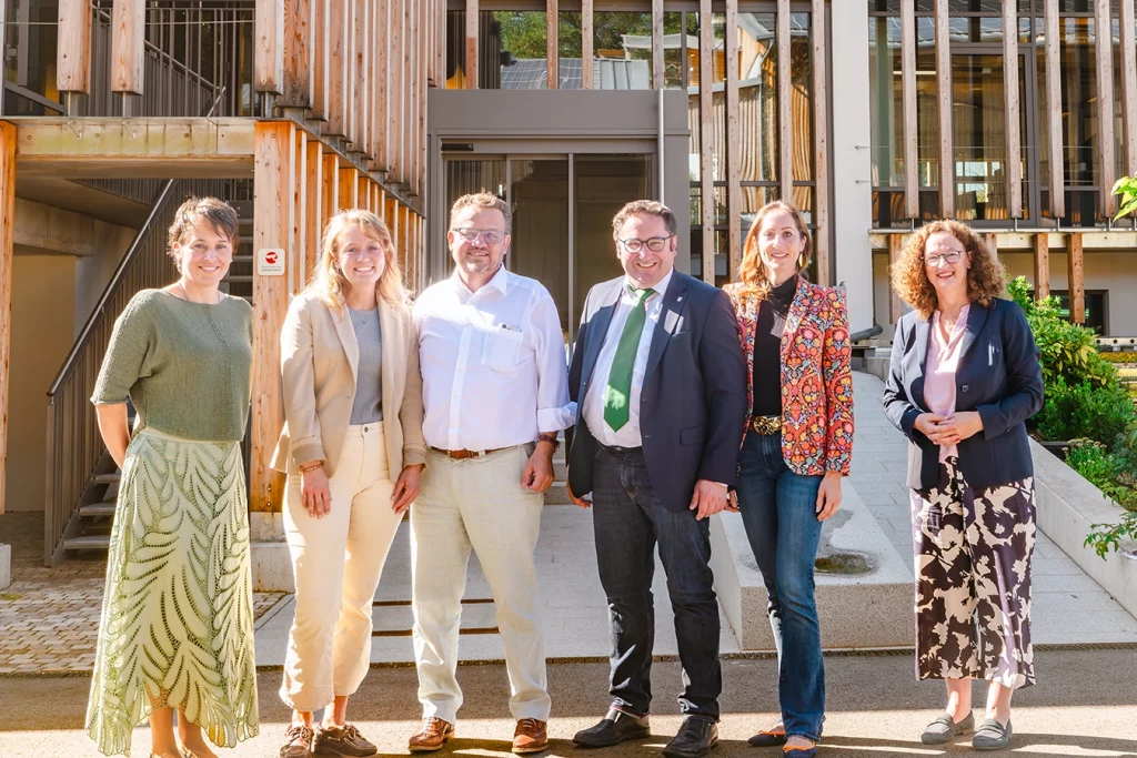 Eine Gruppe von sechs Personen steht vor dem Verwaltungsgebäude der Schmidmeier NaturEnergie. Ganz links steht die Mitarbeiterin Annette Hundsrucker, daneben die Marketingleitern Theresa Schmidmeier. Neben ihr steht der Geschäftsführer Thomas Schmidmeier und anschließend daran der Staatssekretär Tobias Gotthard.