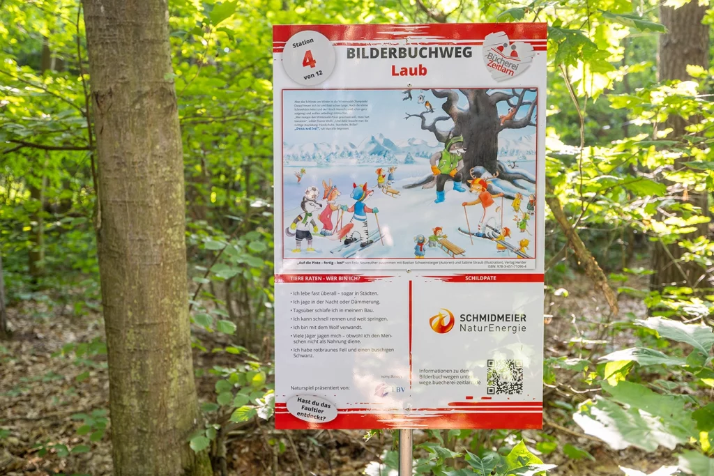 Zu sehen ist ein Schild, welches im Wald angebracht ist. Es gehört zu dem Bilderbuchweg in Zeitlarn. Auf dem Schild gibt es ein Bild sehen mit einem zusammenhängenden Text. welcher beschreibt, was man an dieser Station machen muss. Das Logo von der Schmidmeier ist auf dem Schild platziert, da sie der Schildpate ist.