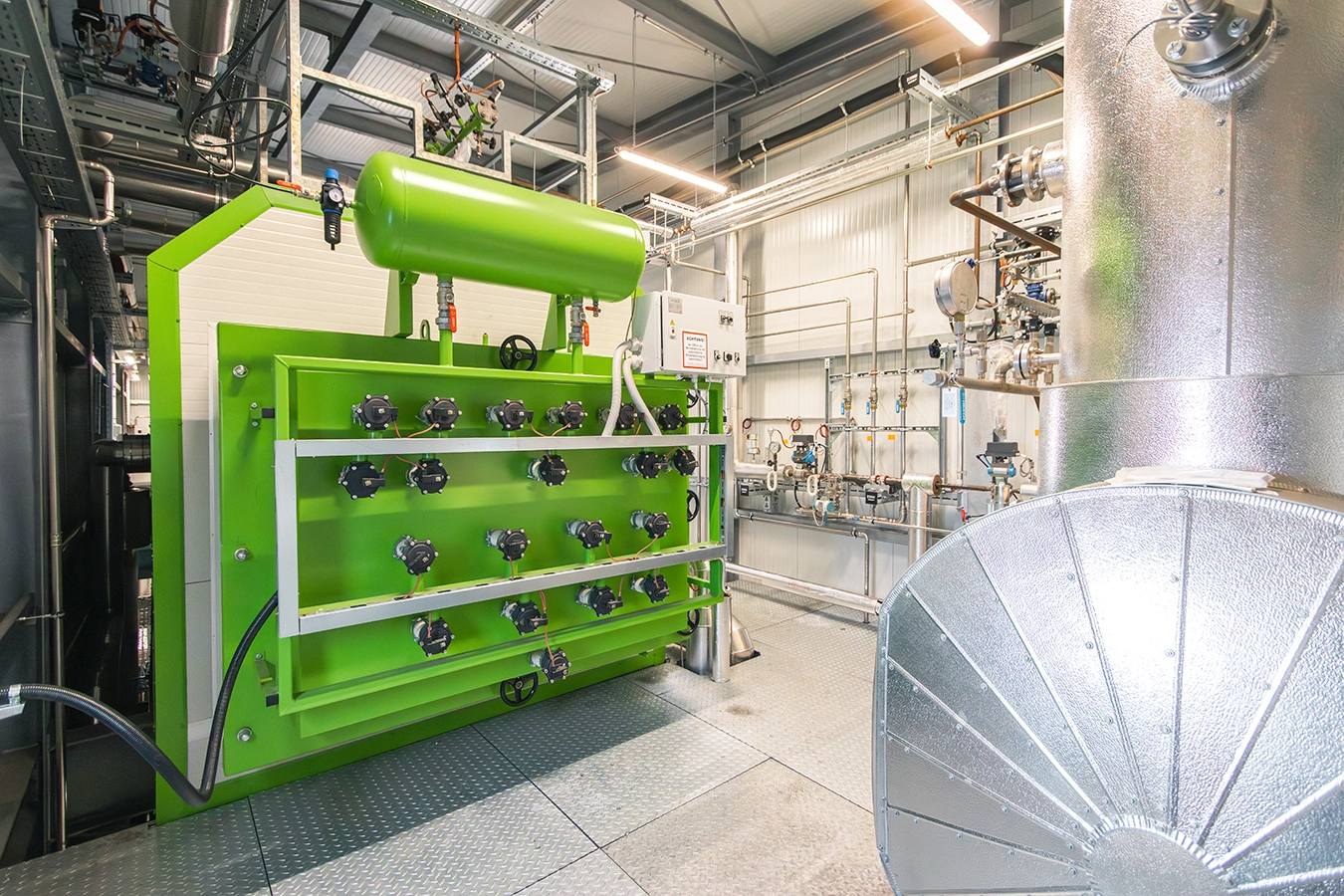 In einem Raum befindet sich ein Teil eines Biomassekessel. Er hat die Farbe Grün. Man erkennt verschiedenste Metallkonstruktionen die Kessel und Rohre Stabilität verleihen.