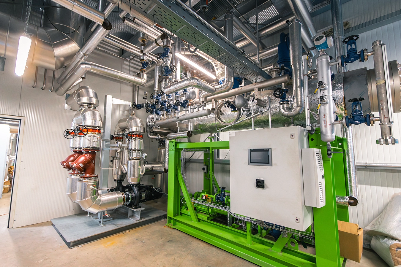 In einem Raum befindet sich ein Teil eines Biomassekessel. Er hat die Farbe Grün. Man erkennt verschiedenste Metallkonstruktionen die Kessel und Rohre Stabilität verleihen.