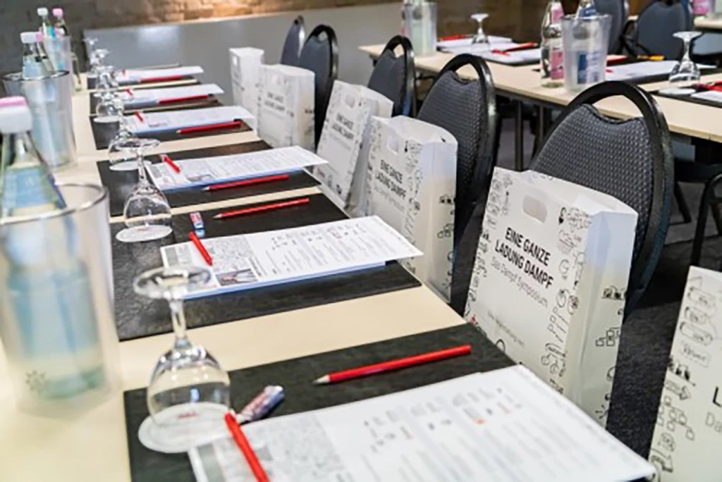 Sitzplätze für die einzelnen Teilnehmer. Auf den Stühlen sind kleine Tüten, auf den Tischen Getränke, Gläser, Stifte und Papier