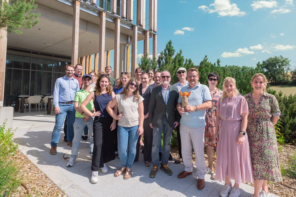Gruppenbild auf dem Weg zu der Terrasse des Verwaltungsgebäude der Schmidmeier NaturEnergie in Zeitlarn. Hinter der Gruppe befinden sich Pflanzen. Der Himmel ist blau, die Sonne scheint.