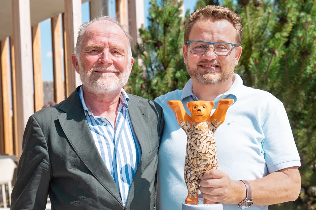 Martin Bentele, Geschäftsführer des Deutschen Energieholz- und Pellet-Verbands, steht neben Thomas Schmidmeier, Geschäftsführer der Schmidmeier NaturEnergie. Sie stehen Arm in Arm. Thomas Schmidmeier hält einen orangen Bären mit Pellets in der Hand. Hinter ihnen ist das Verwaltungsgebäude.
