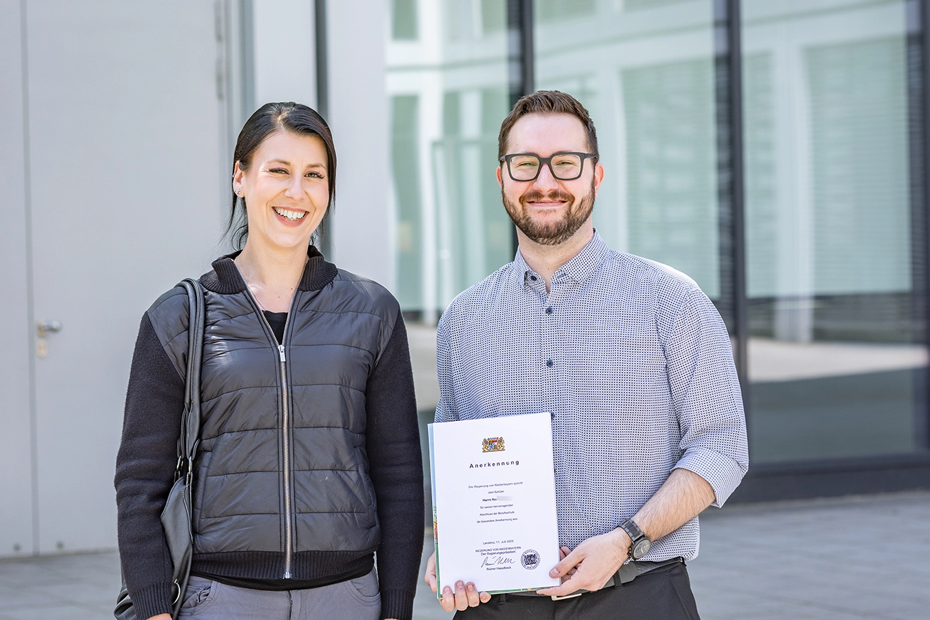Zwei Mitarbeiter der Schmidmeier NaturEnergie stehen lachend nebeneinander. Links, eine Frau und rechts, ein Mann. Der Mann hält eine Urkunde mit der Aufschrift "Anerkennung" in der Hand.