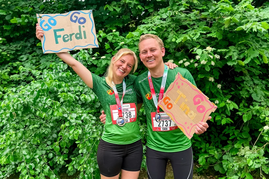 Kristina (links) und Ferdinand Schmidmeier (rechts) stehen in Sportkleidung (grünes Shirt, schwarze Hose) nebeneinander und umarmen sich. Mit ihrer freien Hand halten sie jeweils ein Schild mit den Aufschriften "Go Ferdi Go" und "Go Tina Go". Beide lachen. Sie stehen vor einem grünen Gebüsch.