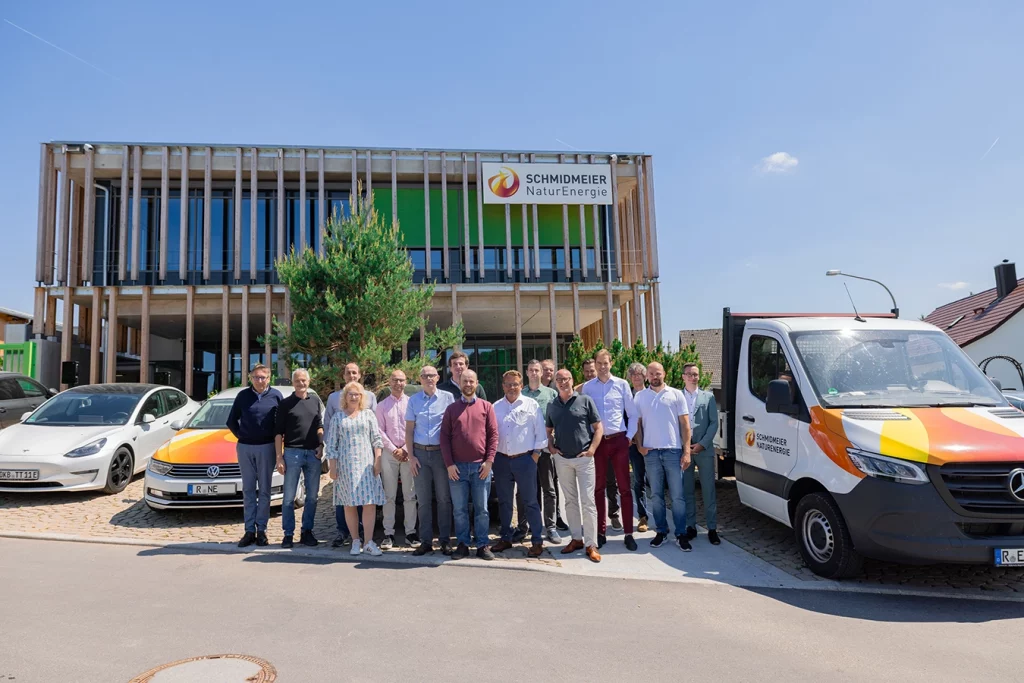 Gruppenbild vor dem Verwaltungsgebäude der Schmidmeier NaturEnergie. Die Personen sind Teil der Fachgruppe Holzheizkraftwerke. Der Himmel ist klar und die Sonne scheint. Neben und hinter der Gruppe befinden sich Dienstwägen von Schmidmeier NaturEnergie, in weiß und orange.
