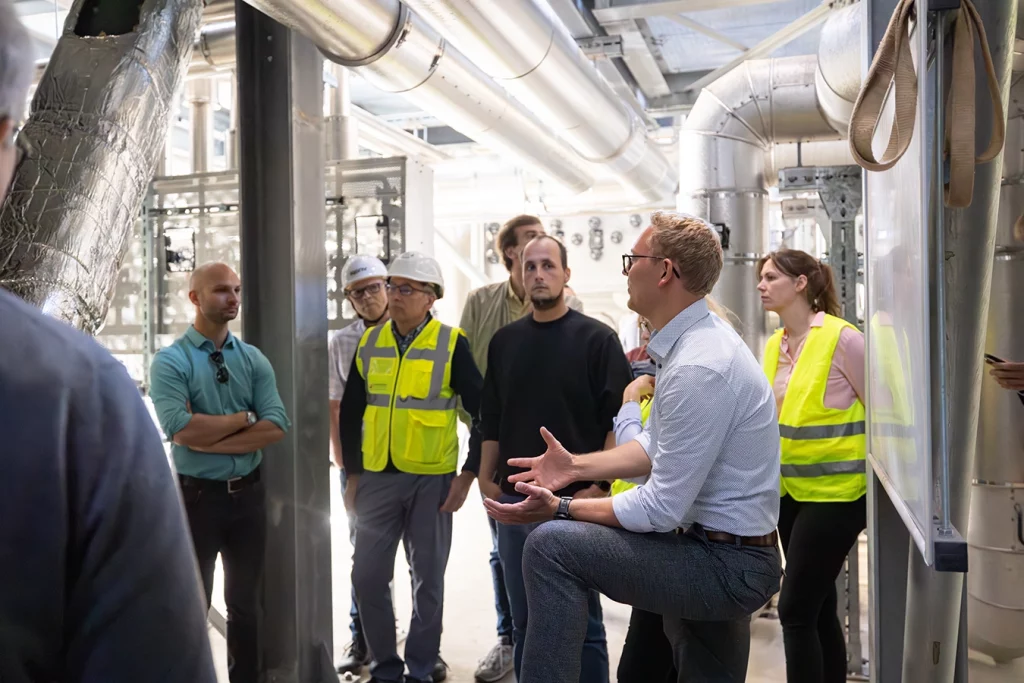 Geschäftsführer Ferdinand Schmidmeier steht innerhalb einer Biomasseanlage. Um ihn herum stehen die Teilnehmer der Führung und hören zu, was Ferdinand Schmidmeier erklärt. Er benutzt dazu seine Hände und stützt dabei die linke Hand auf seinem Knie ab.