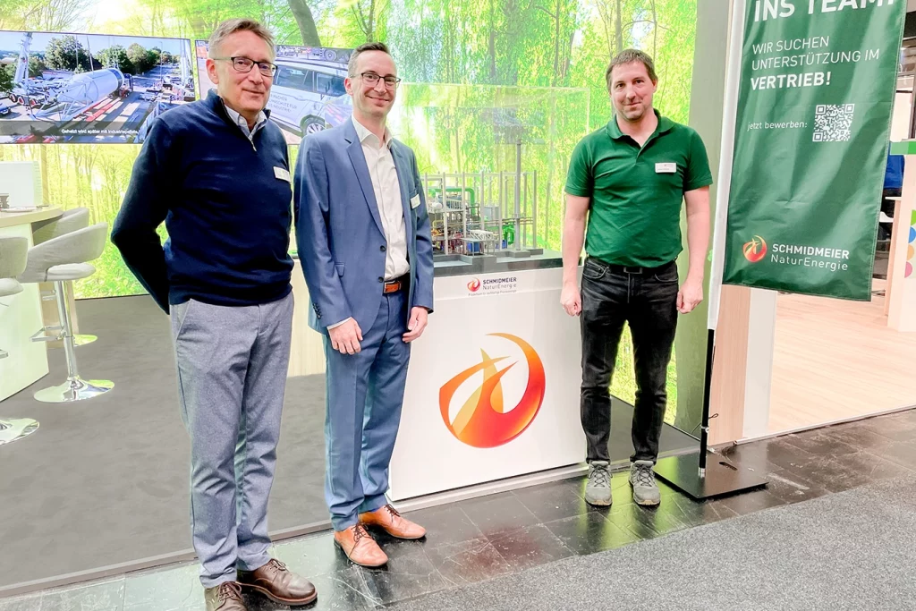 Drei Mitarbeiter der Schmidmeier Natur Energie stehen vor einem Modell einer Biomasseanlage bei ihrem Stand auf der Ligna in Hannover. Links befindet sich Pete Heuer, daneben steht Gerrit Kettel.