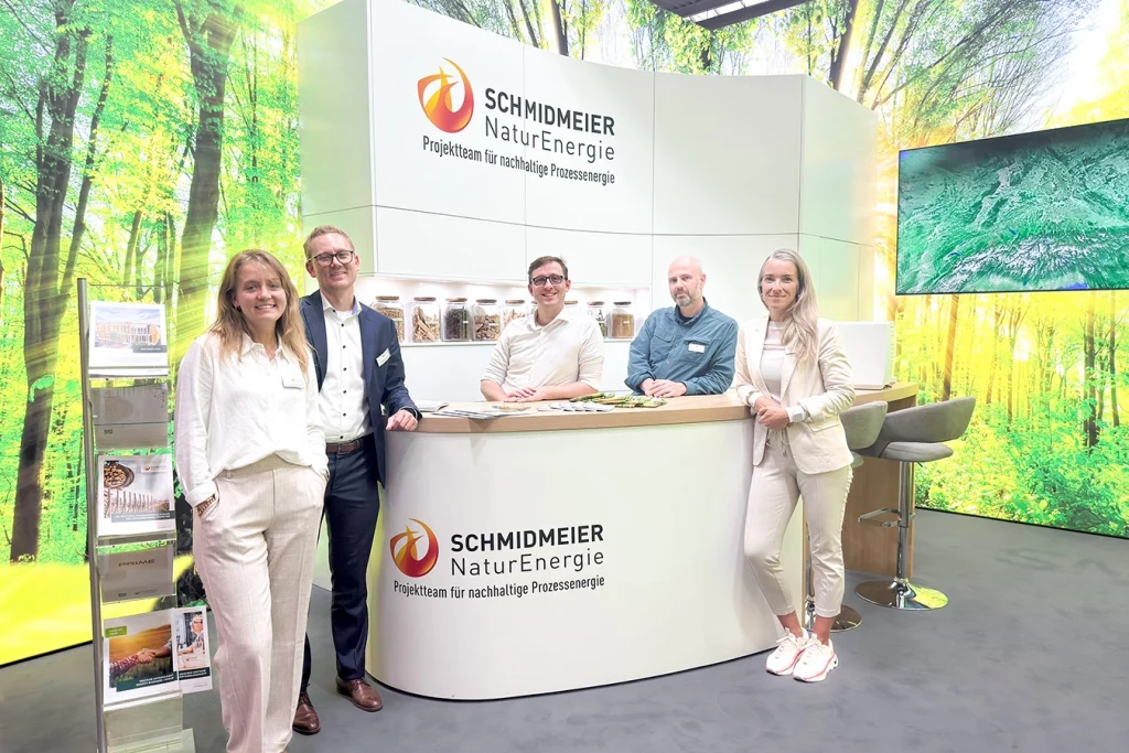 Fünf Mitarbeiter der Schmidmeier NaturEnergie stehen am Messestand auf der Ligna in Hannover. Ganz links im Bild ist Theresa Schmidmeier, Marketingleiterin. Rechts neben ihr steht ihr Bruder Ferdinand Schmidmeier, Geschäftsführer. Ein weißer Tisch sowie Schrank mit Logo der Schmidmeier NaturEnergie befinden sich vor einer Wand, die einen grünen Wald mit hellen Strahlen zeigt.
