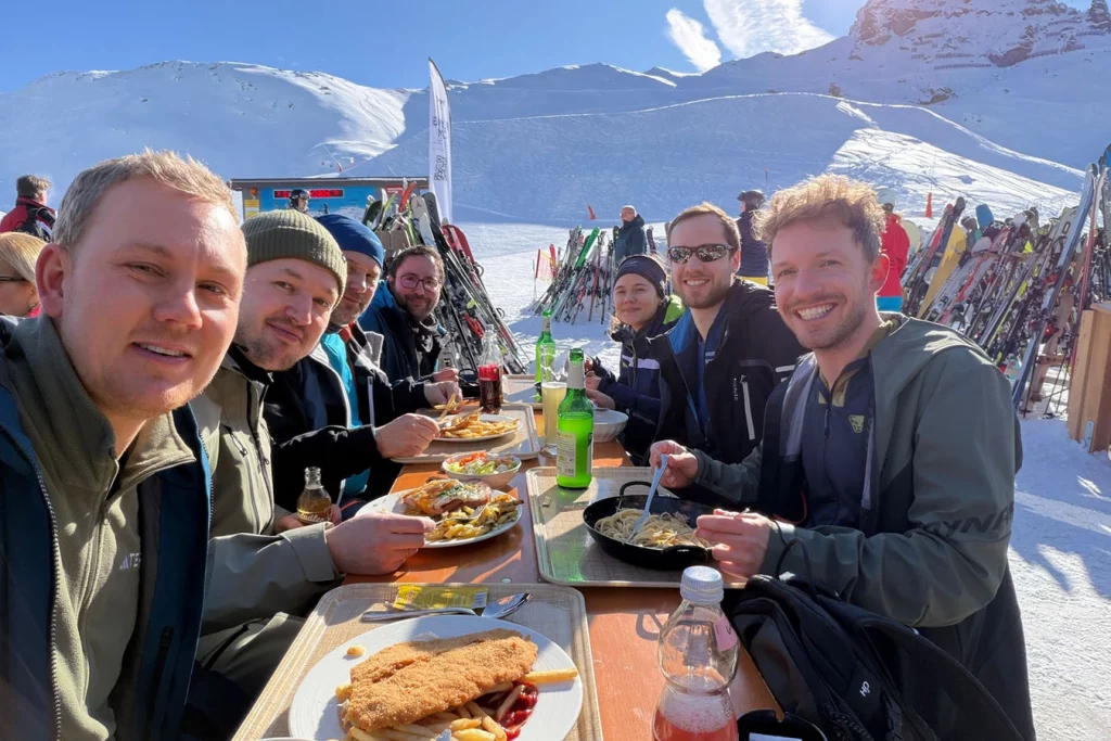 Gruppenbild des Teams beim Essen. Die Sonne scheint und im Hintergrund sieht man die Berge und den Schnee.
