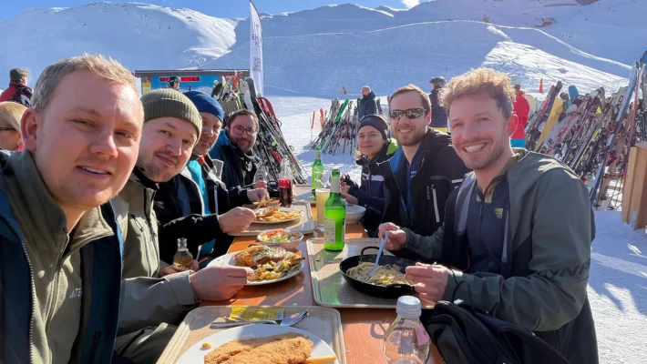 Gruppenbild des Teams beim Essen. Die Sonne scheint und im Hintergrund sieht man die Berge und den Schnee.