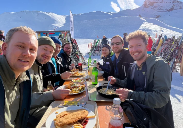 Gruppenbild des Teams beim Essen. Die Sonne scheint und im Hintergrund sieht man die Berge und den Schnee.