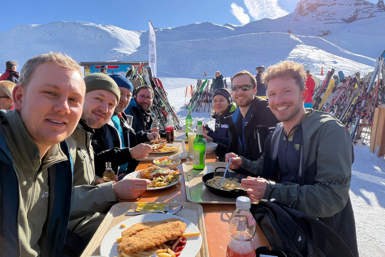 Gruppenbild des Teams beim Essen. Die Sonne scheint und im Hintergrund sieht man die Berge und den Schnee.