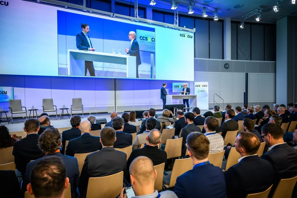 Zwei Personen stehen auf der Bühne der Veranstaltung Handelsblatt Jahrestagung und diskutieren. Ihnen hört dabei ein großes Publikum zu.