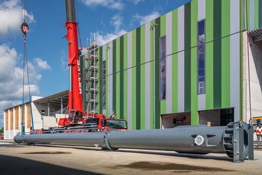 Vor einer Biomasseanlage mit grüner Wand liegt ein grauer Kamin. Der rote LKW, der daneben steht, hat seine Ketten an einem Ende des Kamins.