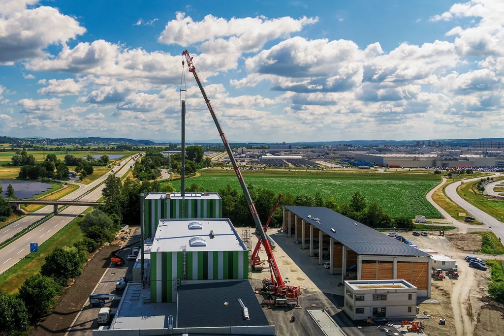 Eine Drohnenaufnahme zeigt die grüne Biomasseanlage in Dingolfing. Ein roter Kran hebt einen Kamin und lässt ihn der Luft schweben. Rechts neben der Biomasseanlage befindet sich eine Lagerhalle, welche teilweise aus Holz gebaut ist.